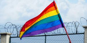 Rainbow flag waving in front of barbed wire under a cloudy sky.