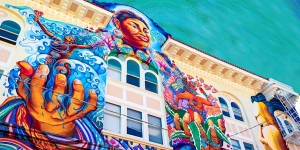 Vibrant mural on a building depicting a woman surrounded by colorful waves and flora, showcasing vivid blues and reds against a clear sky.