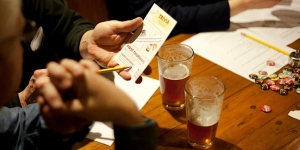 A person holding a trivia card at a table with pint glasses of beer and quiz supplies including pencils and answer sheets.