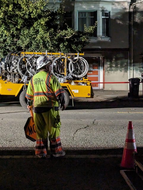 A worker in high visibility clothing stands by the road at night, looking at a truck loaded with bicycles, beside traffic cones.