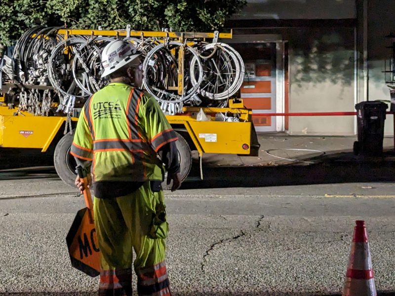 A worker in high visibility clothing stands by the road at night, looking at a truck loaded with bicycles, beside traffic cones.