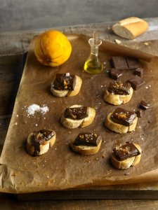 Chocolate and caramel sauce on sliced bread, with sea salt, chocolate bars, and an apple on a wooden table.