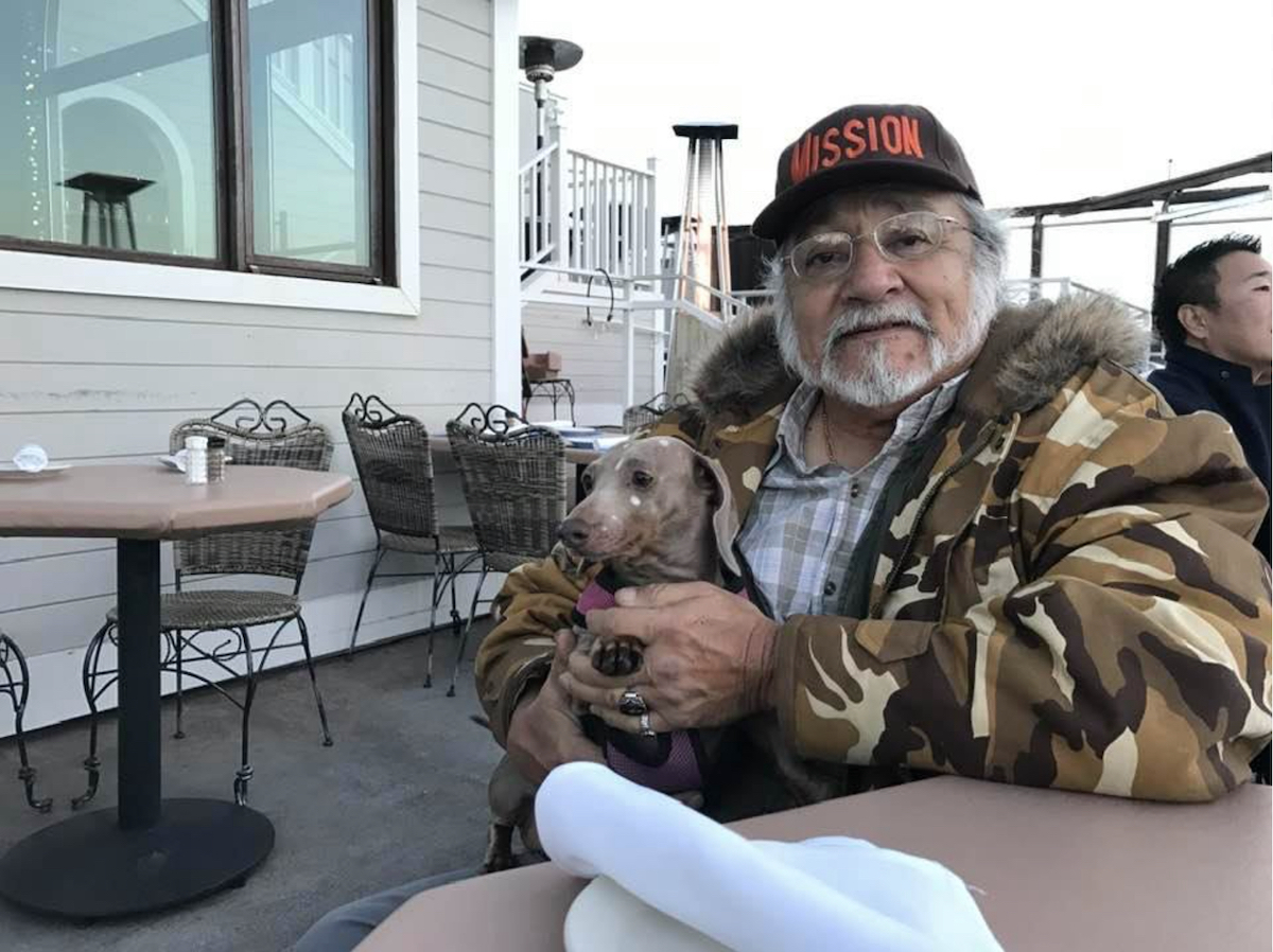 Elderly man with a beard wearing a camouflage jacket and a cap, sitting at an outdoor cafe table holding a small dog.
