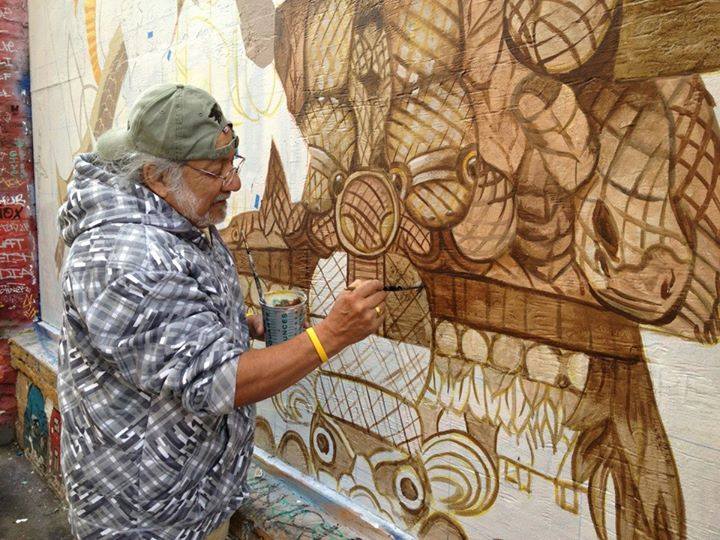An elderly man painting a detailed mural on a wall, using earth tones, focused intently on his work.