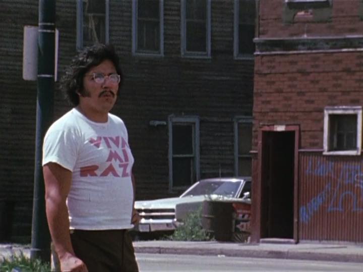 Man with glasses and a white t-shirt stands on a street, with brick buildings and a parked car in the background.
