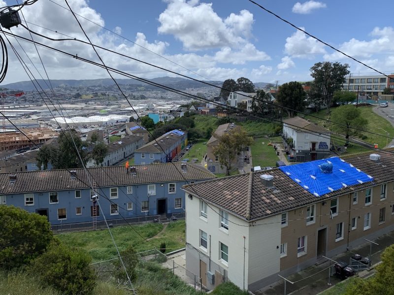Urban slope: a view from above of residential buildings with tarped roof, overlooking an industrial area, under a cloudy sky with power lines crisscrossing overhead.