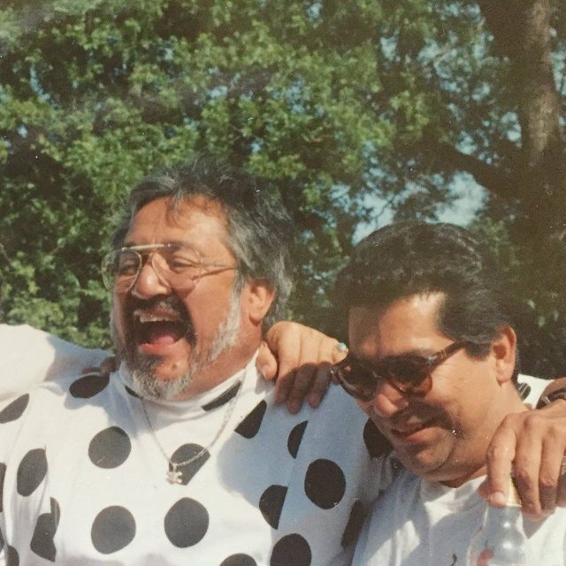 Two middle-aged men smiling outdoors, one wearing a polka dot shirt and glasses, the other in sunglasses, both enjoying a sunny day.