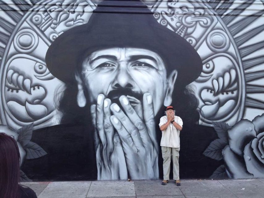A man stands in front of a large mural depicting a detailed, monochromatic portrait of a man with a hat, framed by stylized floral elements.