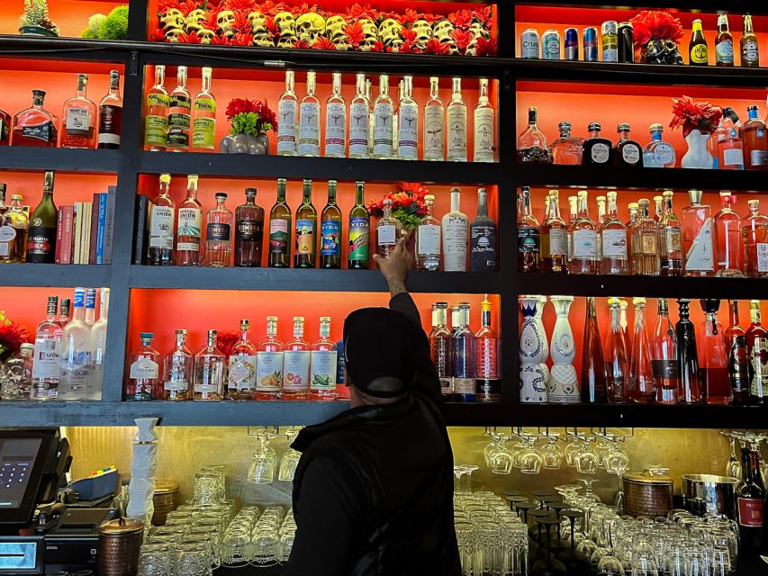 A bartender reaches for a bottle on a shelf filled with various liquors and decorative flowers.