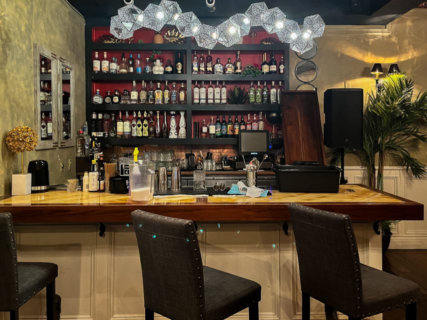 A cozy bar setup with a fully stocked shelf, wooden countertop, two chairs, and decorative lighting.