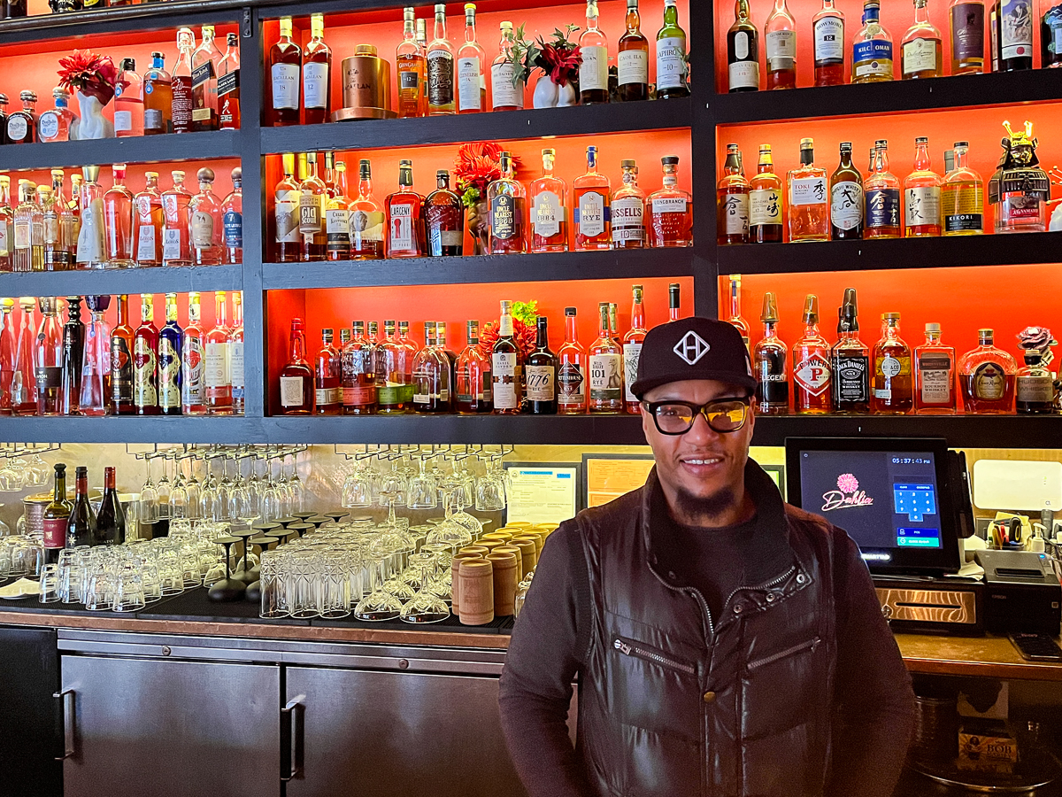 A man smiling in front of a bar with shelves full of various bottles of liquor and wine, glasses neatly arranged below.