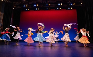 A group of dancers performing on stage in colorful skirts, gracefully waving white scarves in a coordinated dance routine.