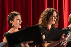 Two women singing with emotion, holding choir folders against a red curtain background.