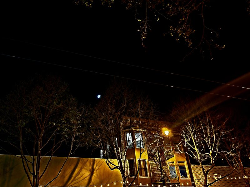 Night-time scene of a warmly lit building, framed by bare trees and utility lines under a dark sky with a bright moon.