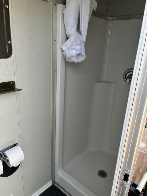 Interior of a compact bathroom featuring a small shower stall with a hanging curtain, toilet paper holder, and a metallic shelf.