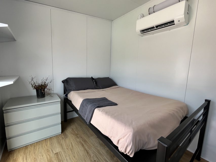 A simple bedroom featuring a double bed with gray and pink bedding, a white dresser, wooden flooring, and an air conditioning unit above the bed.