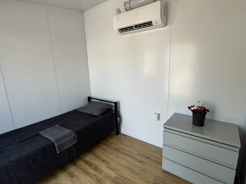 A small, simple bedroom with a single bed, grey bedding, an air conditioning unit above, and a dresser with a red potted plant on it.