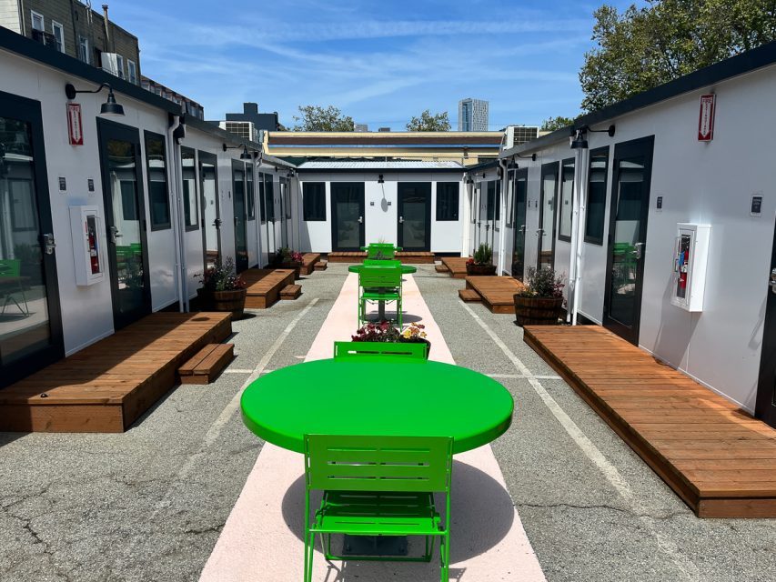 Outdoor seating area between rows of white cabins with green tables and chairs.