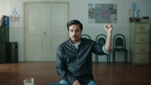 A man sitting on a chair in a classroom, raising his hand as if asking a question, with a world map and cabinet in the background.