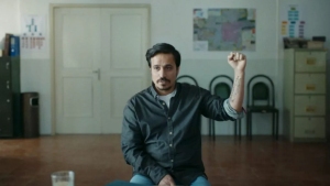 A man sits in a room with his right arm raised, showing a tattoo. The room has a door, chairs, a map on the wall, and a row of filing drawers.