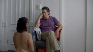 A man in a purple shirt sits on a stool, attentively listening to a woman in a white towel who sits facing him in a minimally furnished room.