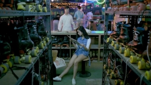 A woman sits on a stool reading a magazine in a roller skate rental shop, surrounded by shelves filled with skates. A few people stand in the background near neon signs.