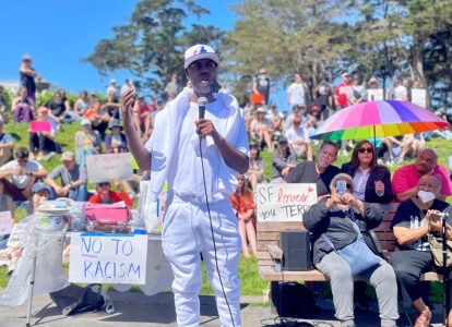 Terry Williams speaking into a microphone at an anti-racism rally in a park, with audience members holding signs and a colorful umbrella visible in the background.