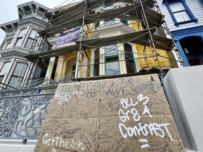 A yellow house under renovation with scaffolding, next to a cardboard sign covered in handwritten messages.