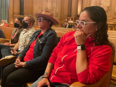 Two people wearing masks and casual clothing seated in a decorative hall, attentively listening to a speaker out of view.