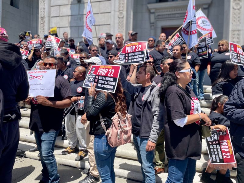 A group of people on building steps holding signs and banners in support of Proposition 22.
