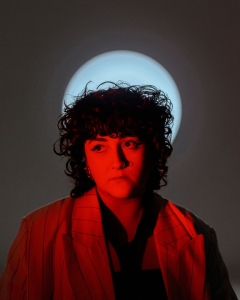 A person with curly hair is illuminated by red light, standing in front of a bright circular light source. They are wearing a striped blazer and looking slightly to the side.