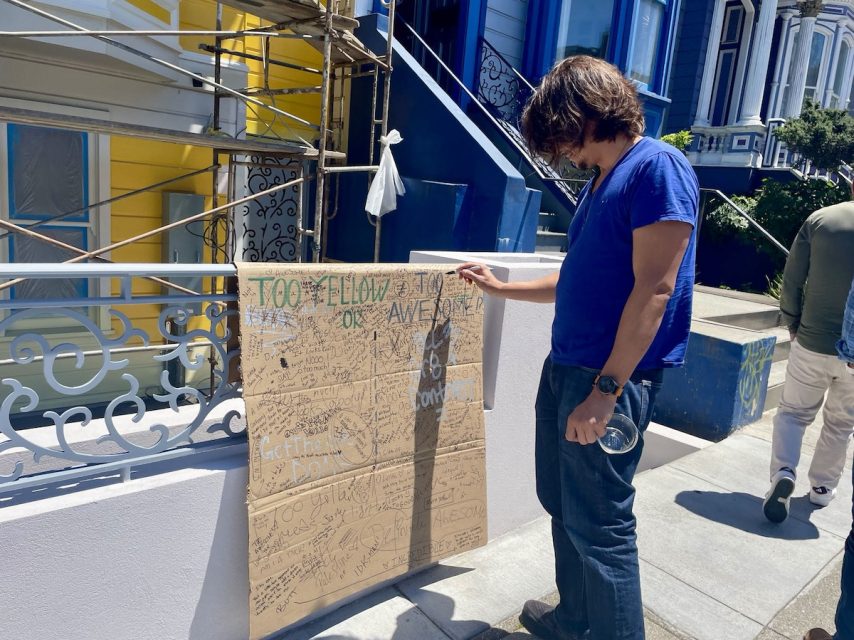 A person stands on the sidewalk, drawing on a large cardboard sign propped against a railing. The sign has multiple doodles and messages. Scaffolding and colorful buildings are in the background.