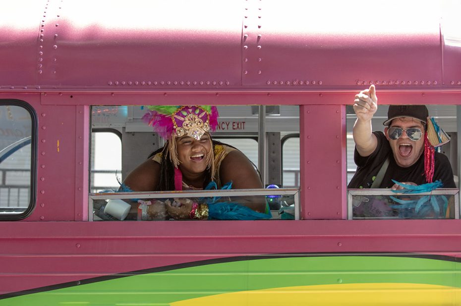 Two people wearing colorful festive costumes and accessories are joyfully leaning out of the windows of a brightly painted bus.