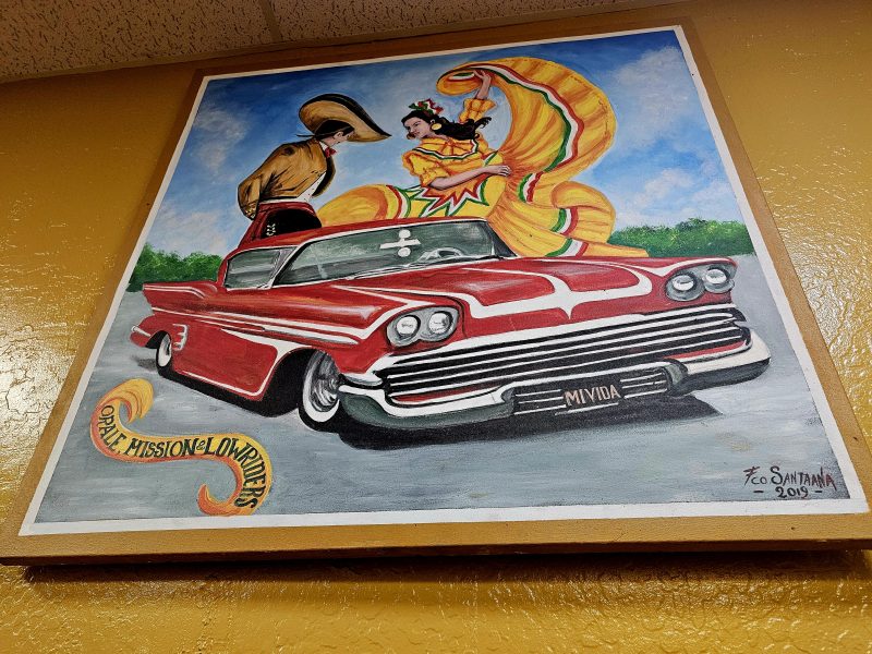 Colorful painting of a classic red lowrier car topped with a man in a cowboy hat and a woman in a flowing yellow dress dancing, framed on a yellow wall.