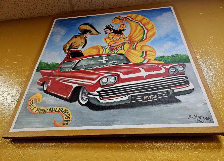 Colorful painting of a classic red lowrier car topped with a man in a cowboy hat and a woman in a flowing yellow dress dancing, framed on a yellow wall.