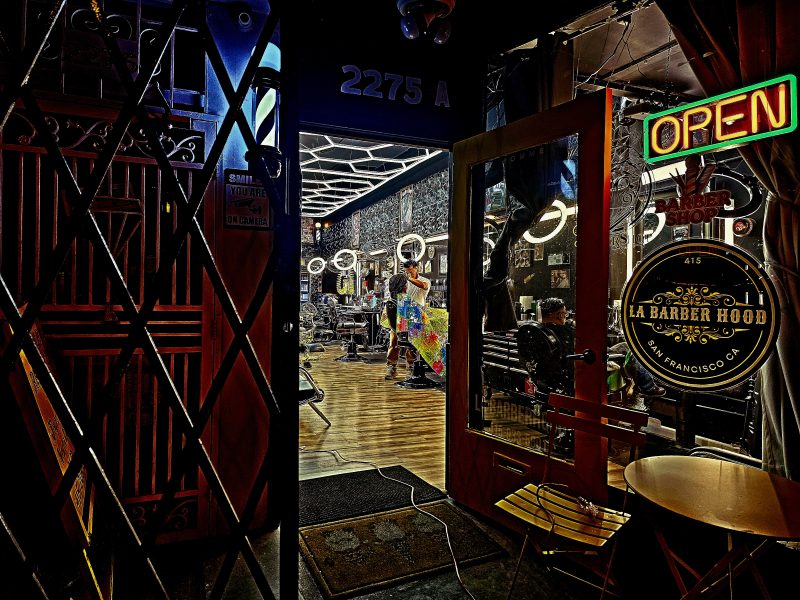 A barber shop at night with an open door, neon "OPEN" sign, and lit interior showing a barber working on a client. The exterior has a grid security gate and the address 2275 A.