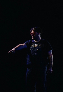 A person wearing sunglasses and dark clothing stands in a dark environment, pointing with their right hand.