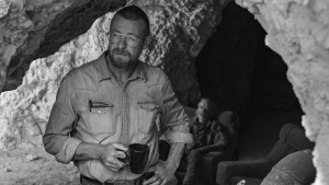 A man with glasses and a beard, wearing a shirt and holding a mug, stands inside a cave with another person seated in the background. black and white photo.