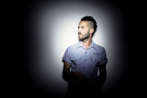 A man with a beard and tattoos, wearing a short-sleeve shirt, stands against a plain, dark background, looking to the right.