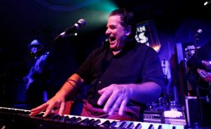 Man playing piano and singing into a microphone on a stage lit with purple lights.