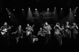 Band performs on stage under spotlights at the "city of django" festival, with members playing various instruments like guitars, violin, and trumpet.