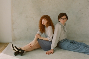 A woman with curly red hair and a man with glasses, both dressed in smart casual outfits, sitting back-to-back on a concrete floor.