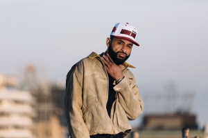 A man in a beige jacket and red baseball cap standing outdoors with his hand on his chin, looking thoughtful.
