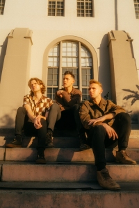 Three young men sitting on steps outside a building with arched windows, bathed in warm sunlight.