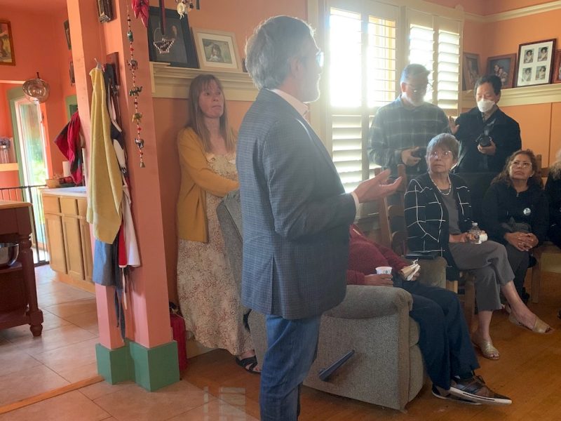 A man in a suit speaks to a small group of seated and standing people in a colorful room with orange walls and various decorations.