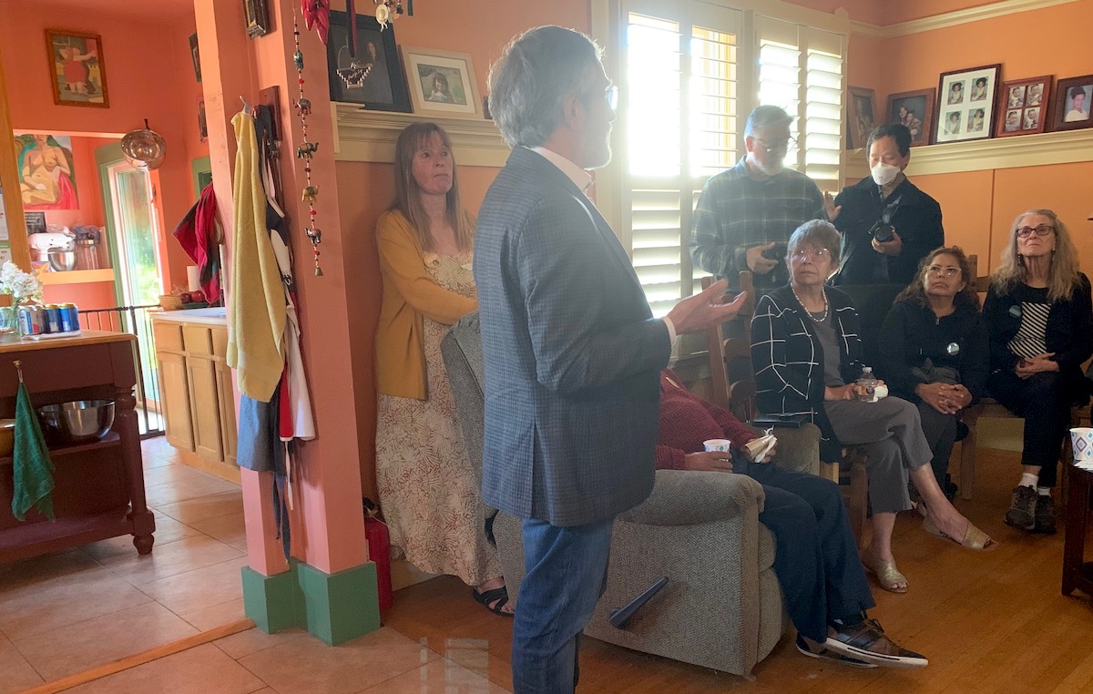 A man in a suit speaks to a small group of seated and standing people in a colorful room with orange walls and various decorations.