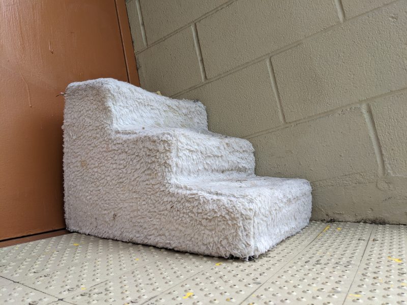 A set of carpeted pet stairs with white fluffy material, placed next to a brown door and against a beige tiled wall.