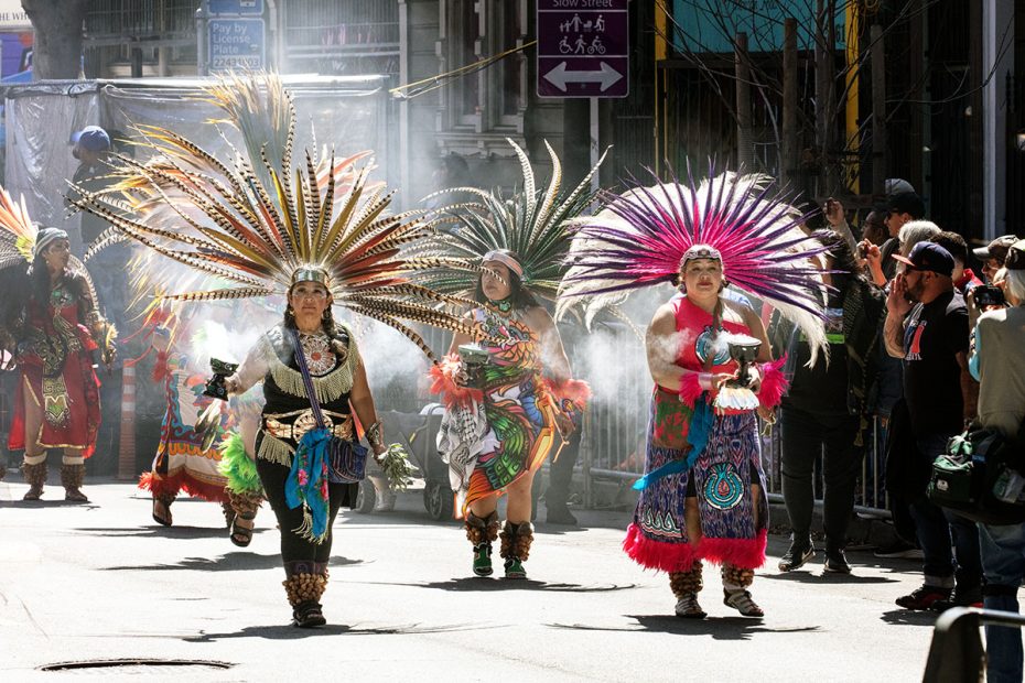 A group of people in colorful, traditional costumes and feathered headdresses perform a dance on a street, surrounded by onlookers and photographers.