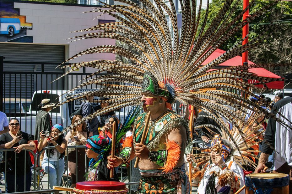 A dancer dressed in traditional attire with a large feathered headdress performs in a street parade, surrounded by onlookers.