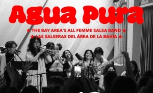 A group of women performing music on stage under a sign that reads "Agua Pura: The Bay Area's All Femme Salsa Band / Las Salseras del Área de la Bahía.